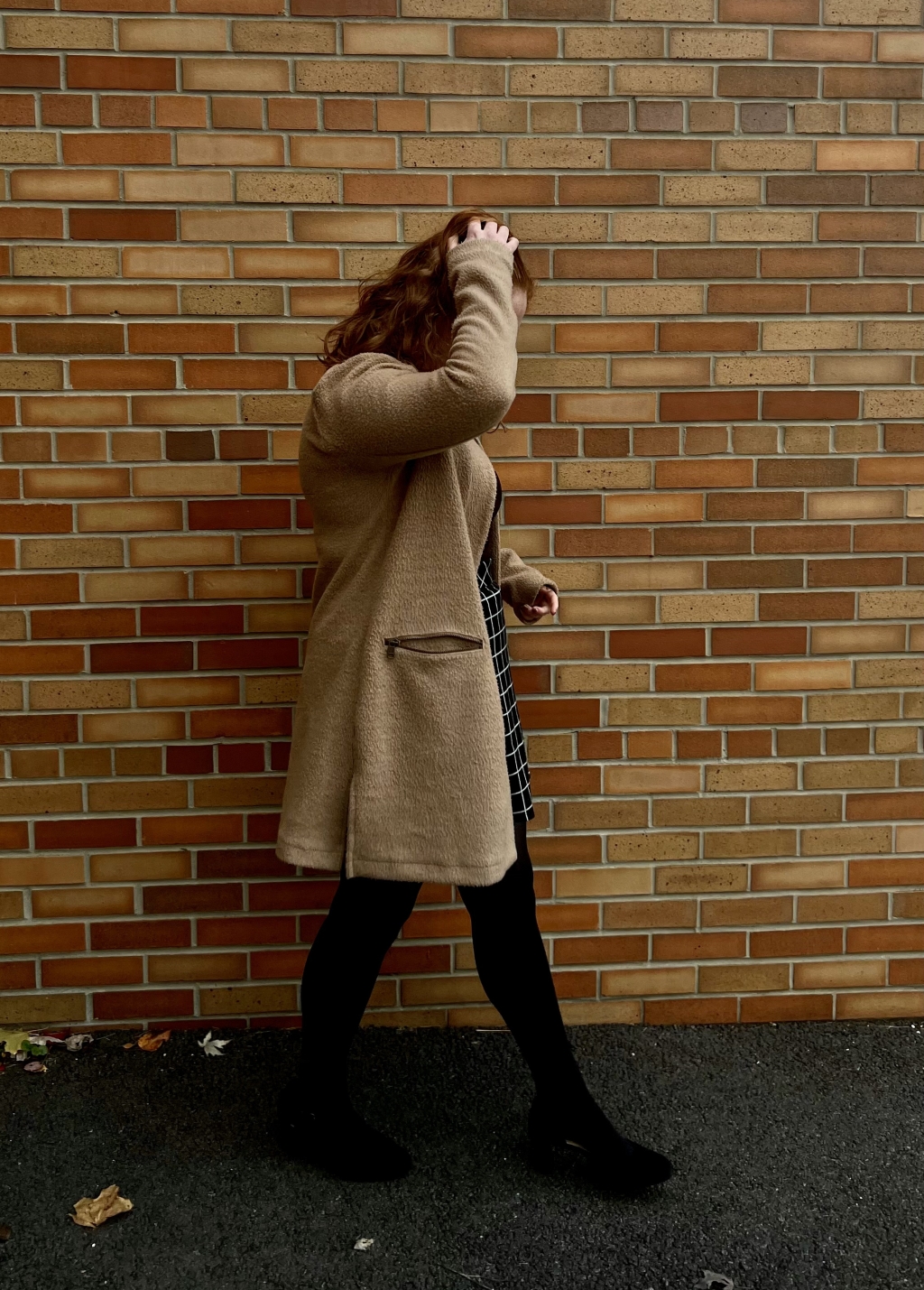 Woman in tan coat looks at ground and walks alongside a brick building wither her hand combing through her hair